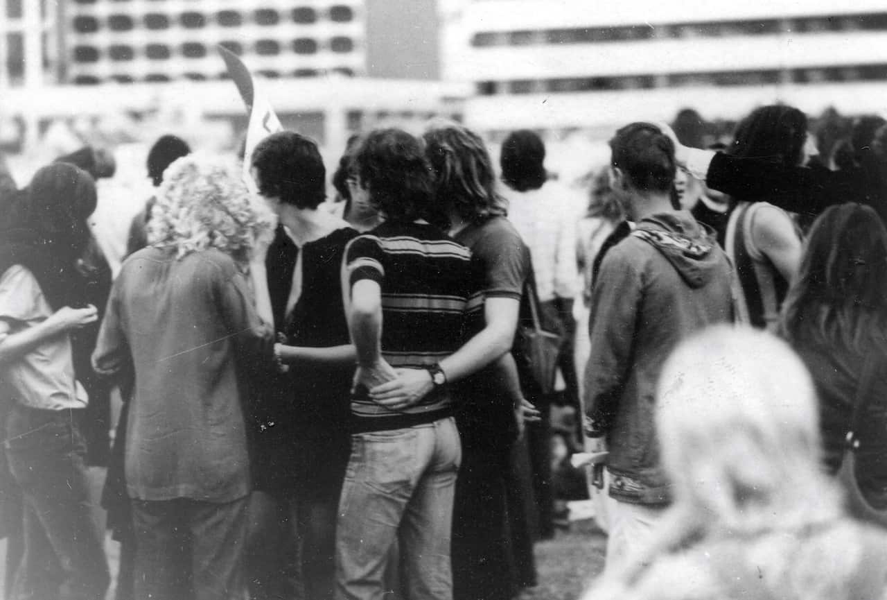 A black and white image of a man with his arm around another man's waist, holding hands. They are in a crowd of people with their backs to the camera.