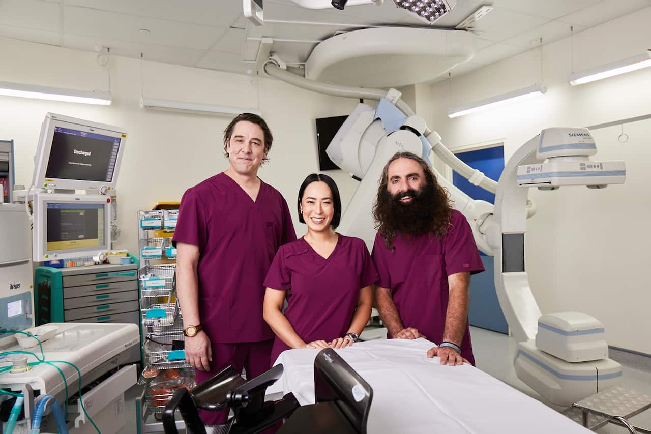 A woman and two men, wearing maroon scrubs, stand in a hospital treatment room. 