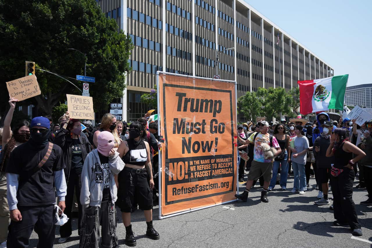 A large group of protesters, some holding up a big orange banner that reads "Trump must go now".