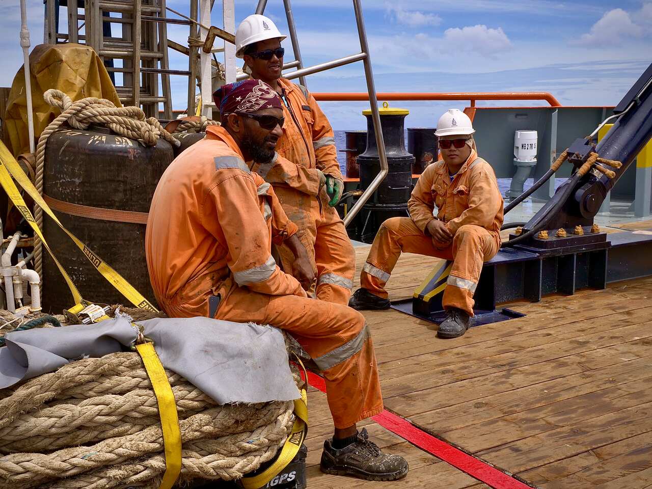 Three crew members sit of the back deck of the ship in orange overalls. 