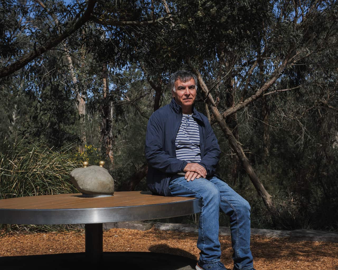 A middle-aged man with dark hair sits on the edge of a large, circular metal table outdoors, surrounded by trees and dry leaves on the ground. He is wearing a dark blue jacket over a striped shirt and jeans, with his hands clasped in his lap.
