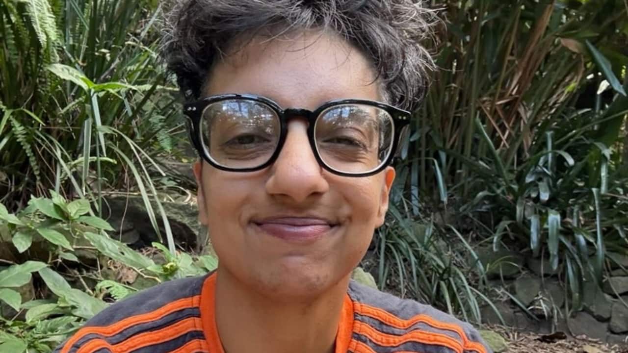 a closeup shot of a person with black framed glasses and in a orange top sits in a garden