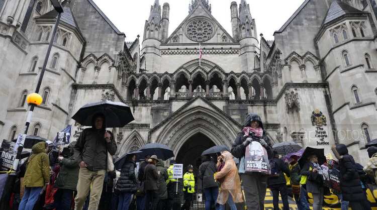 People dressed in down jackets, with several holding umbrellas gather outside a cathedral. Many in the crowd hold signs, with one reading 'Hands of Julian Assange.'