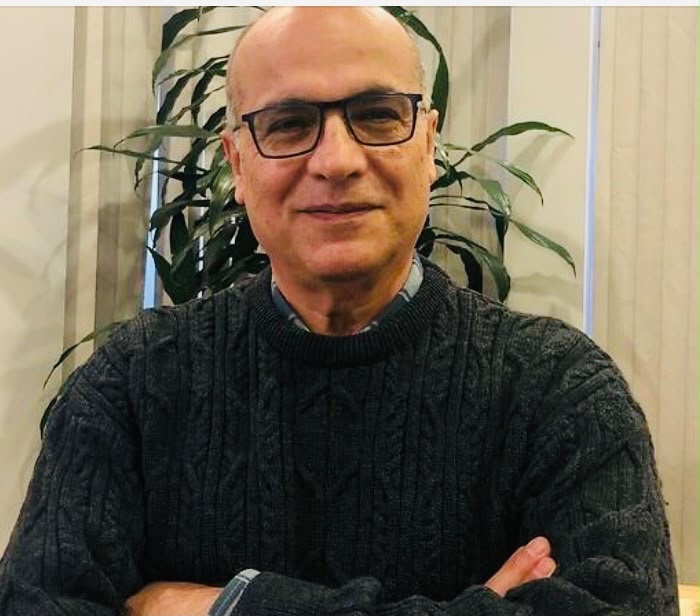 A man in glasses and a black round-neck sweater sits in front of an indoor plant with his arms crossed in front.