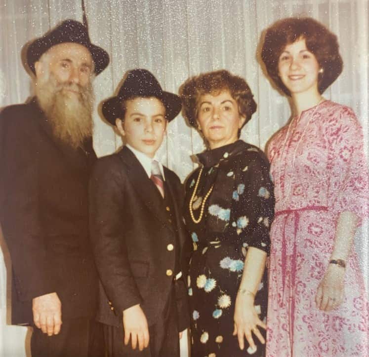 A man with a grey beard stands next to a child weaing a hat, another woman in a black and floral dress and a woman in a pink dress.