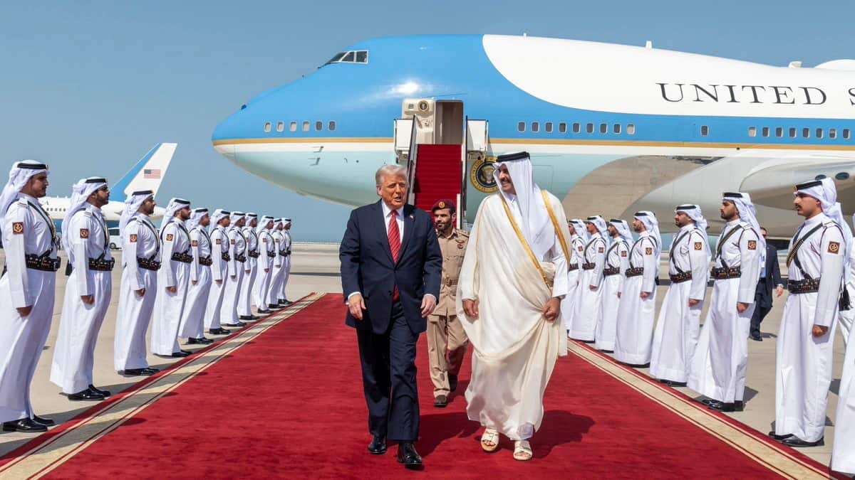 A man in a dark suit walks next to a man in a thobe down a red carpet, with a large blue jet behind them. Qatari Amiri Guard officials in uniforms stand in straight lines on both sides of the carpet.