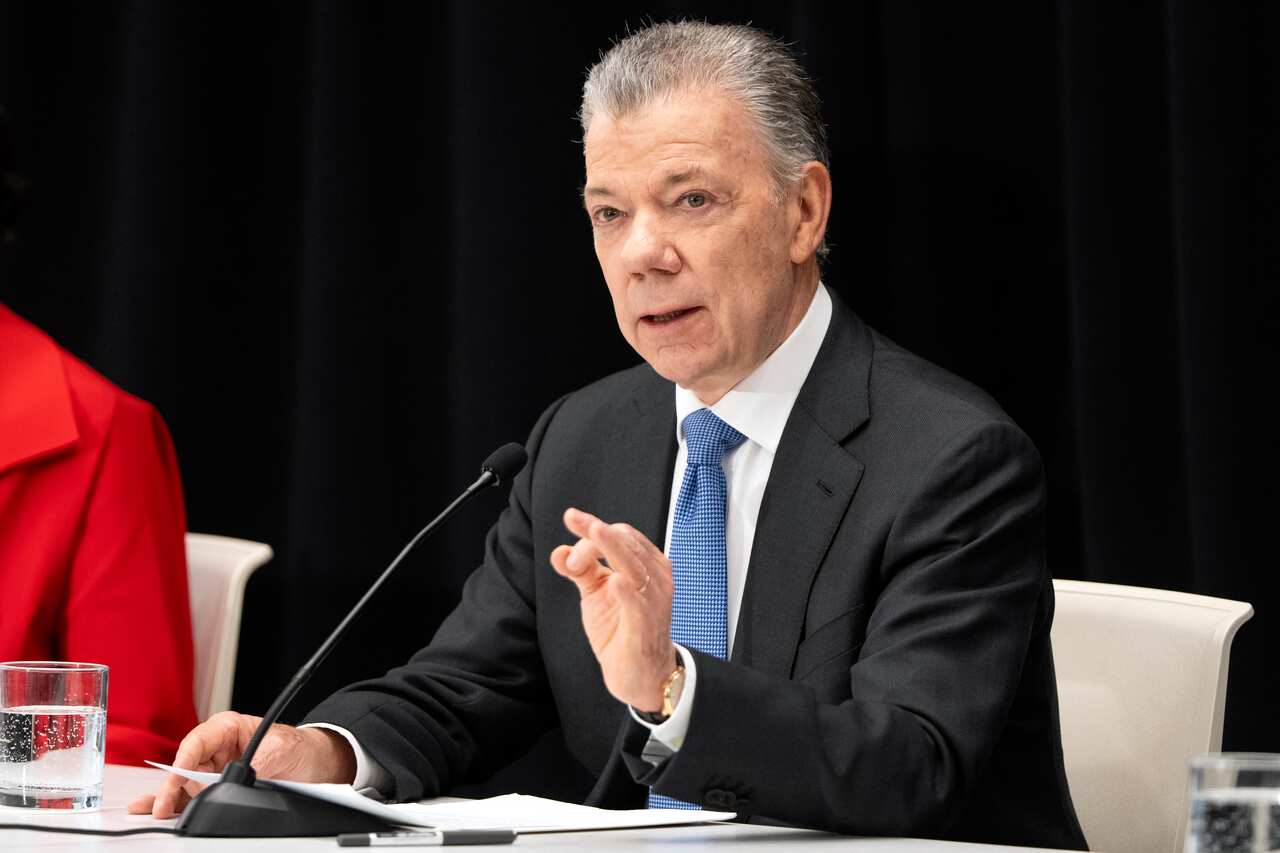 A man in a black suit, white shirt and blue tie sits at a desk and speaks into a microphone.