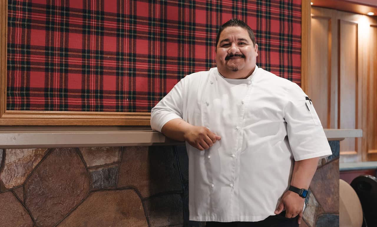 A man in a chef's jackets tands in a room with a tartan wall, leaning on an elbow-height ledge. 