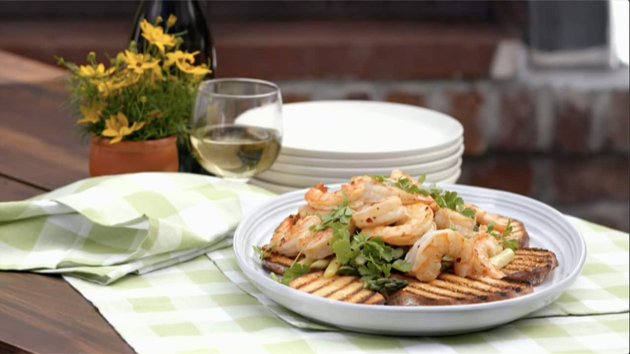 A shallow white bowl holds slices of grilled bread topped with a prawn, asparagus and herb mixture. The bowl sits on a green and white checked tablecloth. Stacked plates, a glass of white wine and a pot with yellow flowers sit behind the bowl. 