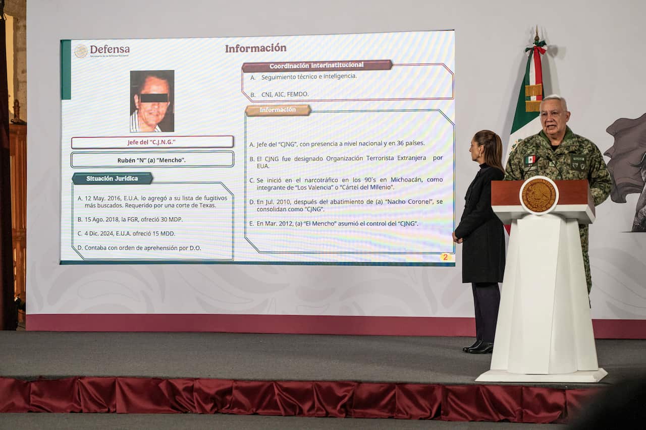 A uniformed Mexican military official speaks at a podium beside a large screen displaying information about a cartel leader during a government press briefing.