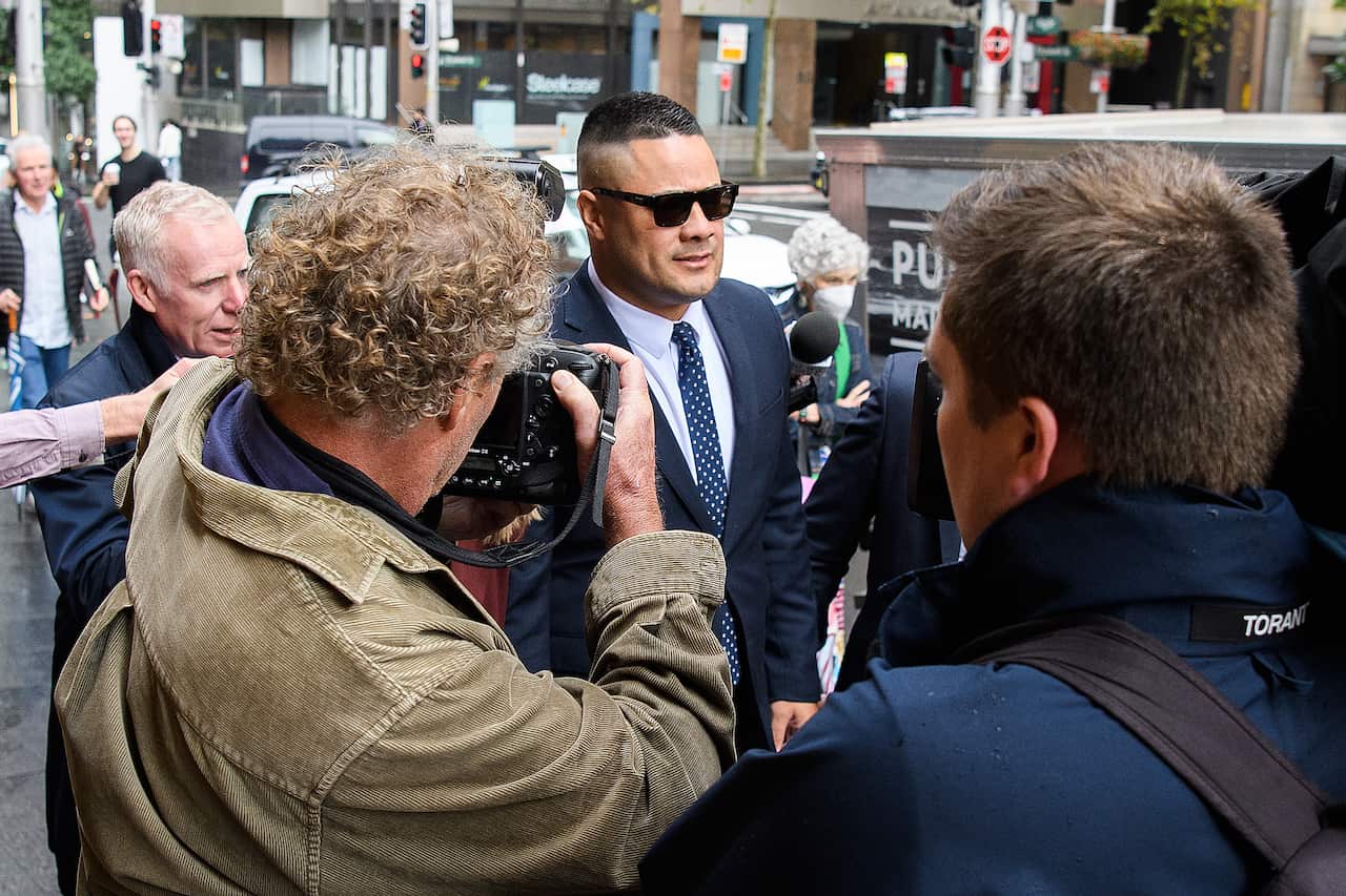 A man wearing sunglasses and a suit is surrounded by media photographers. 