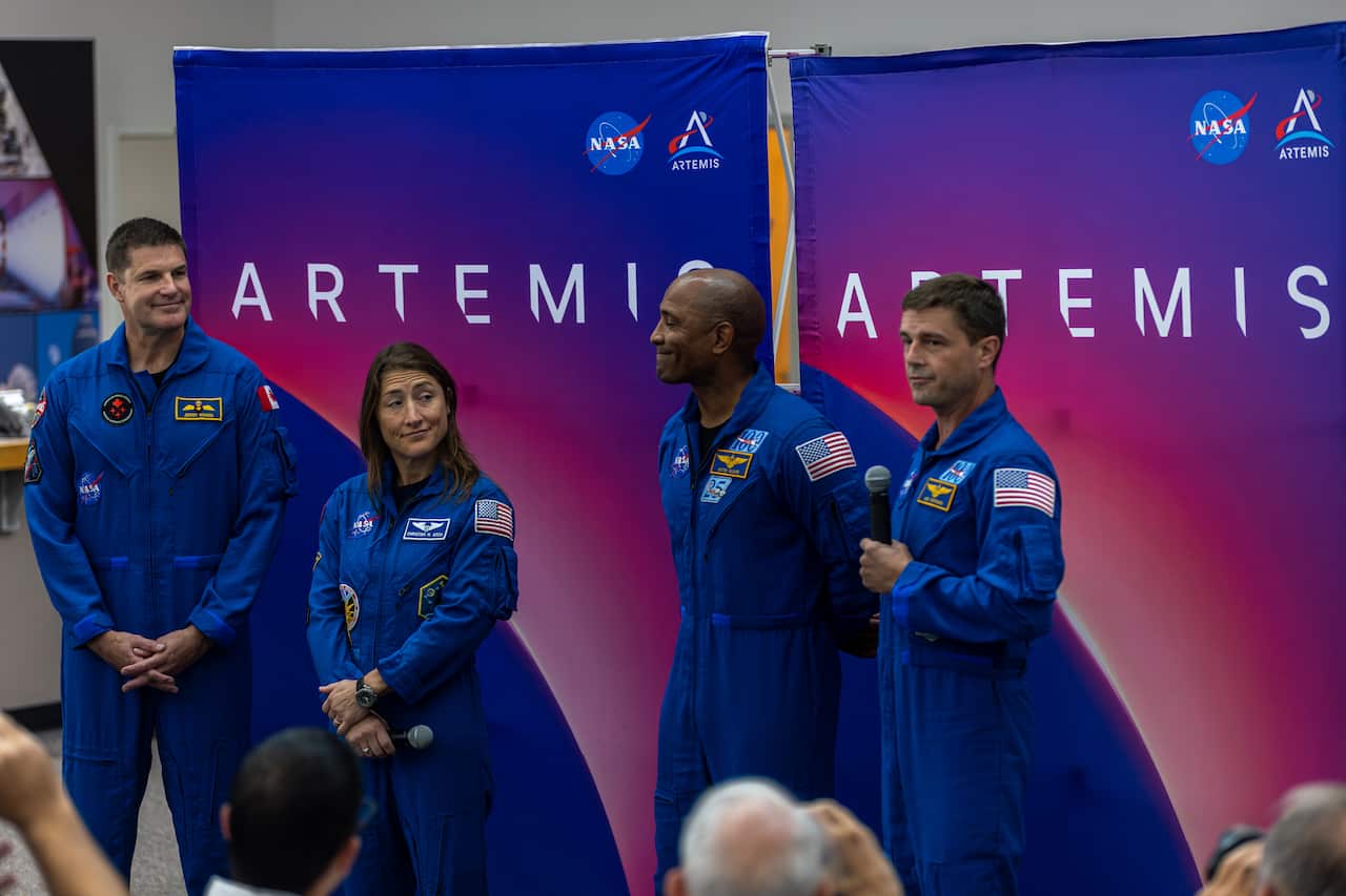 A photograph shows four NASA astronauts standing in a row, all wearing blue flight suits. They are in front of a blue and purple backdrop that says "Artemis."