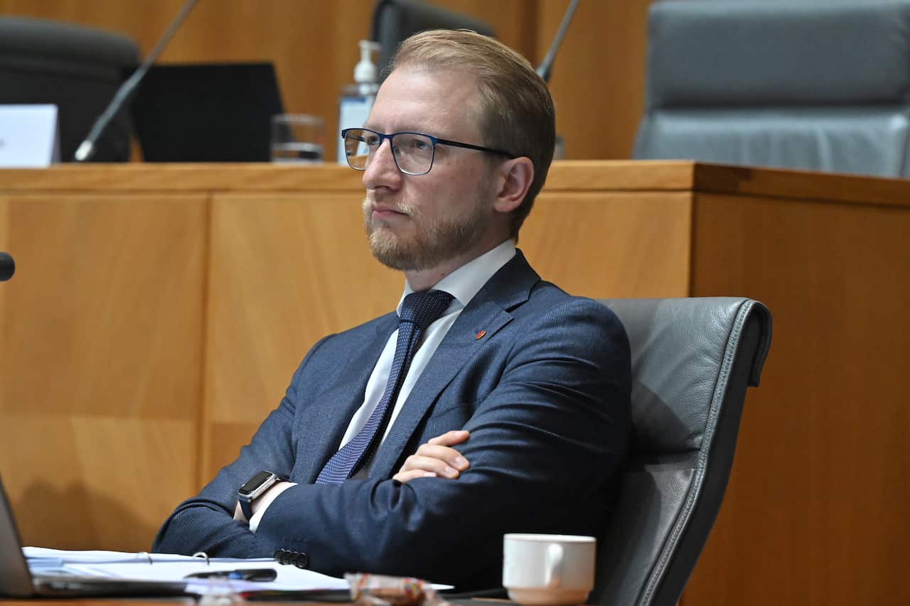A politician wearing glasses and a dark blue suit sits back in a chair with his arms folded.
