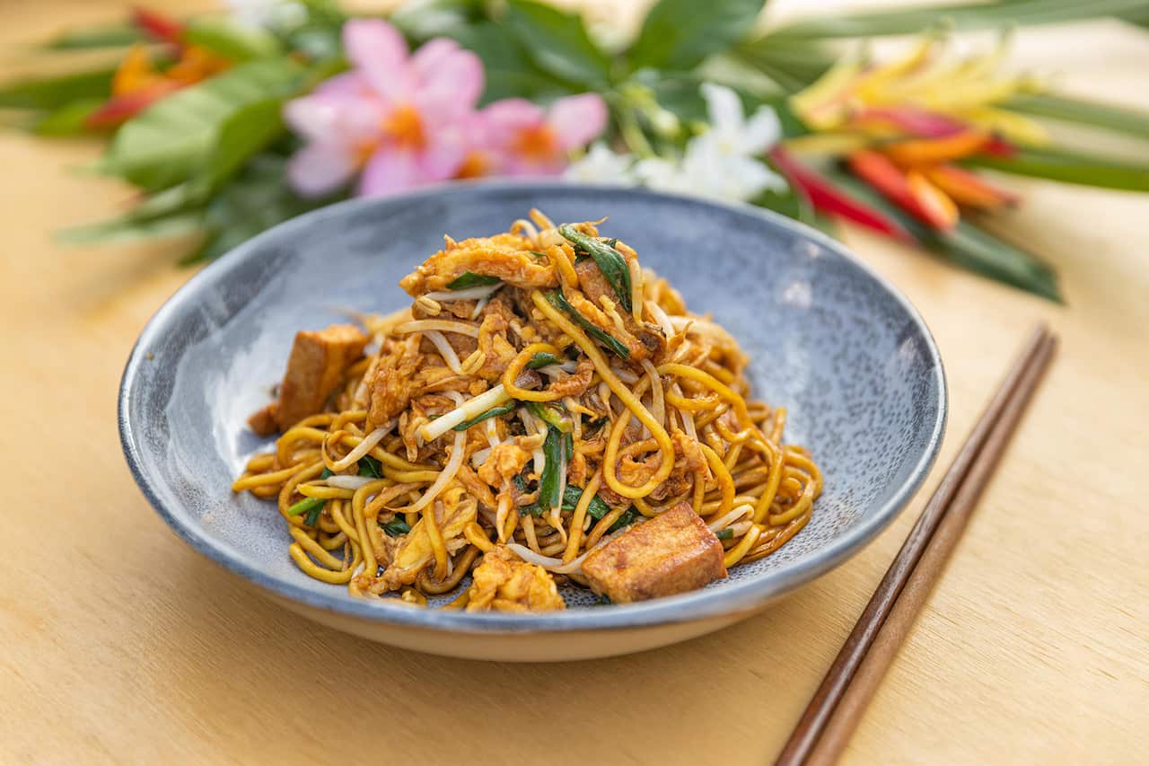 A beautiful blue-grey bowl sits on a wooden surface. It holds a pile of cooked noodles with egg, tofu and greens. A set of chopsticks sits to one side of the bowl. Fresh flowers can be seen in the background. 