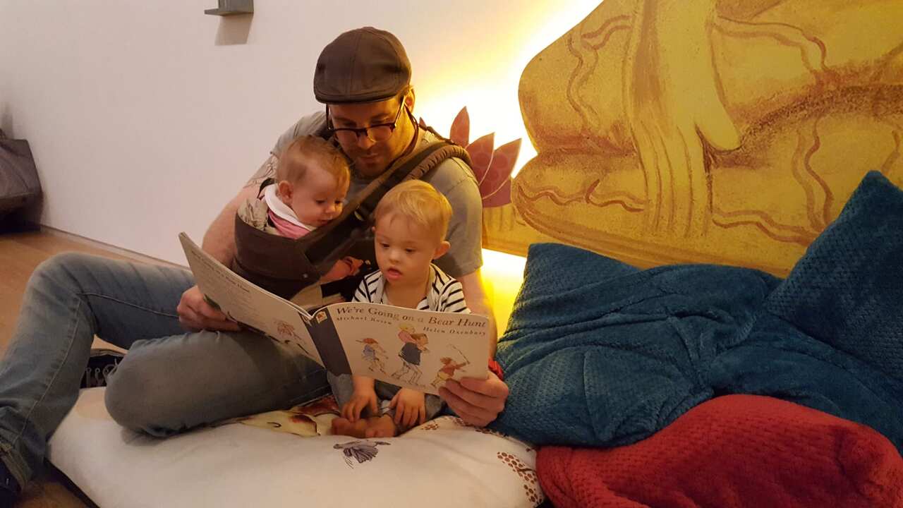 A man with a baby strapped to his body, sits and reads a book to a little boy sitting next to him