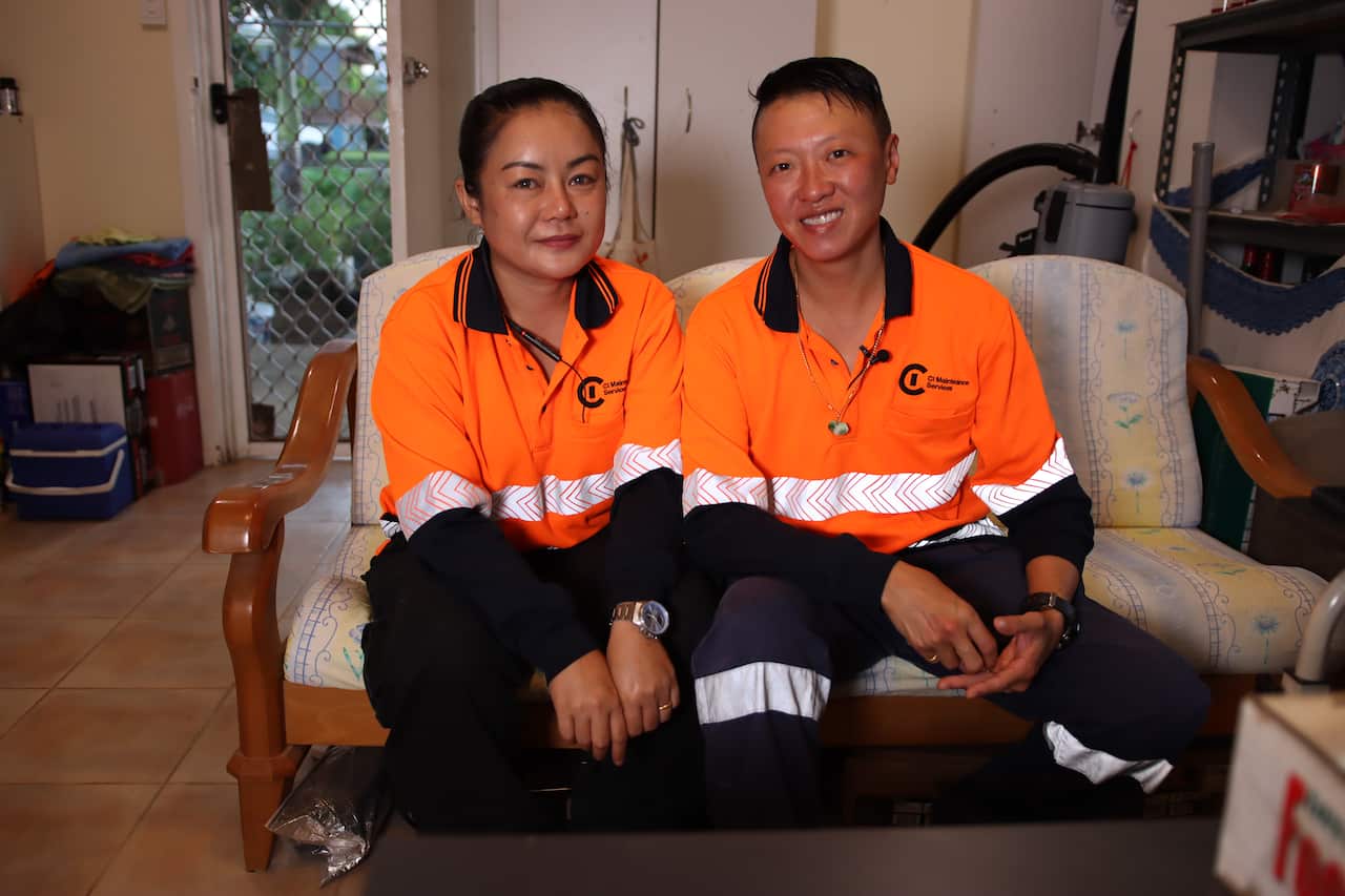 Two women wearing orange hi-vis shirts sit on a couch inside a property on Christmas Island