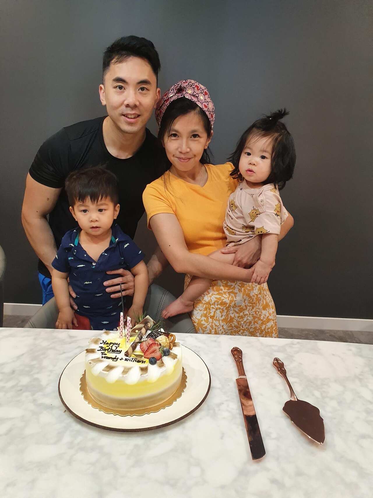A family of four stand behind a birthday cake. The mum and dad are smiling, and the little boy and girl are looking straight ahead.