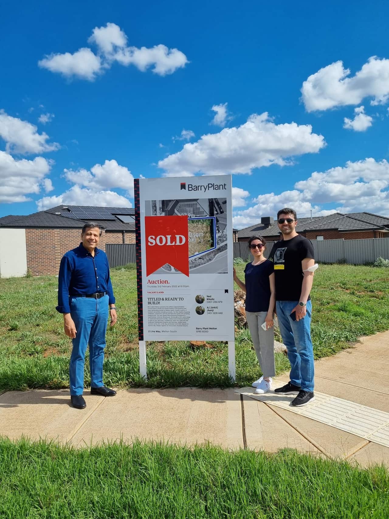 Two men and a woman stand smiling in front of a "SOLD" real estate sign in front of a grassy vacant lot in a suburban neighbourhood with houses visible in the background.