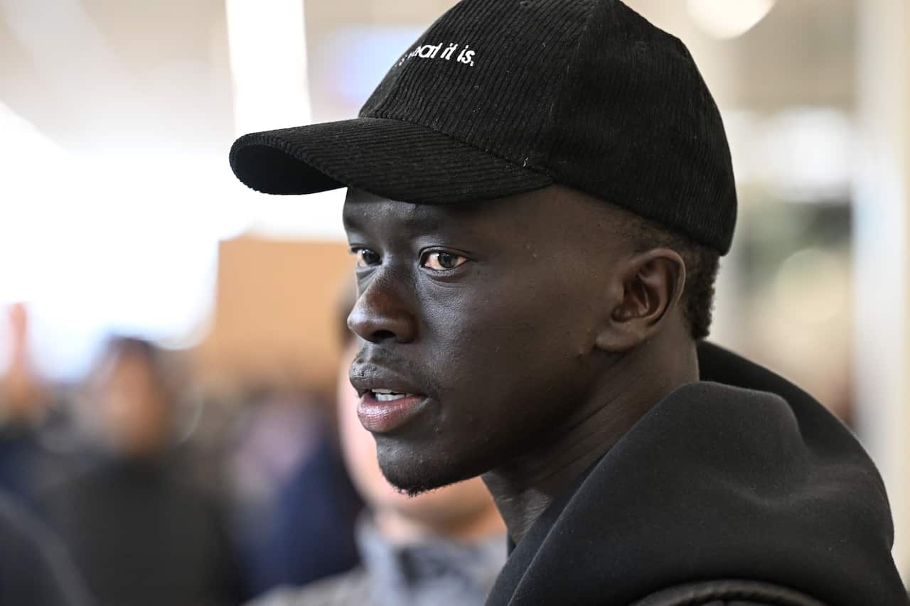 Headshot of Awer Mabil wearing a black hoodie and cap, background is blurred so only his face in focus.