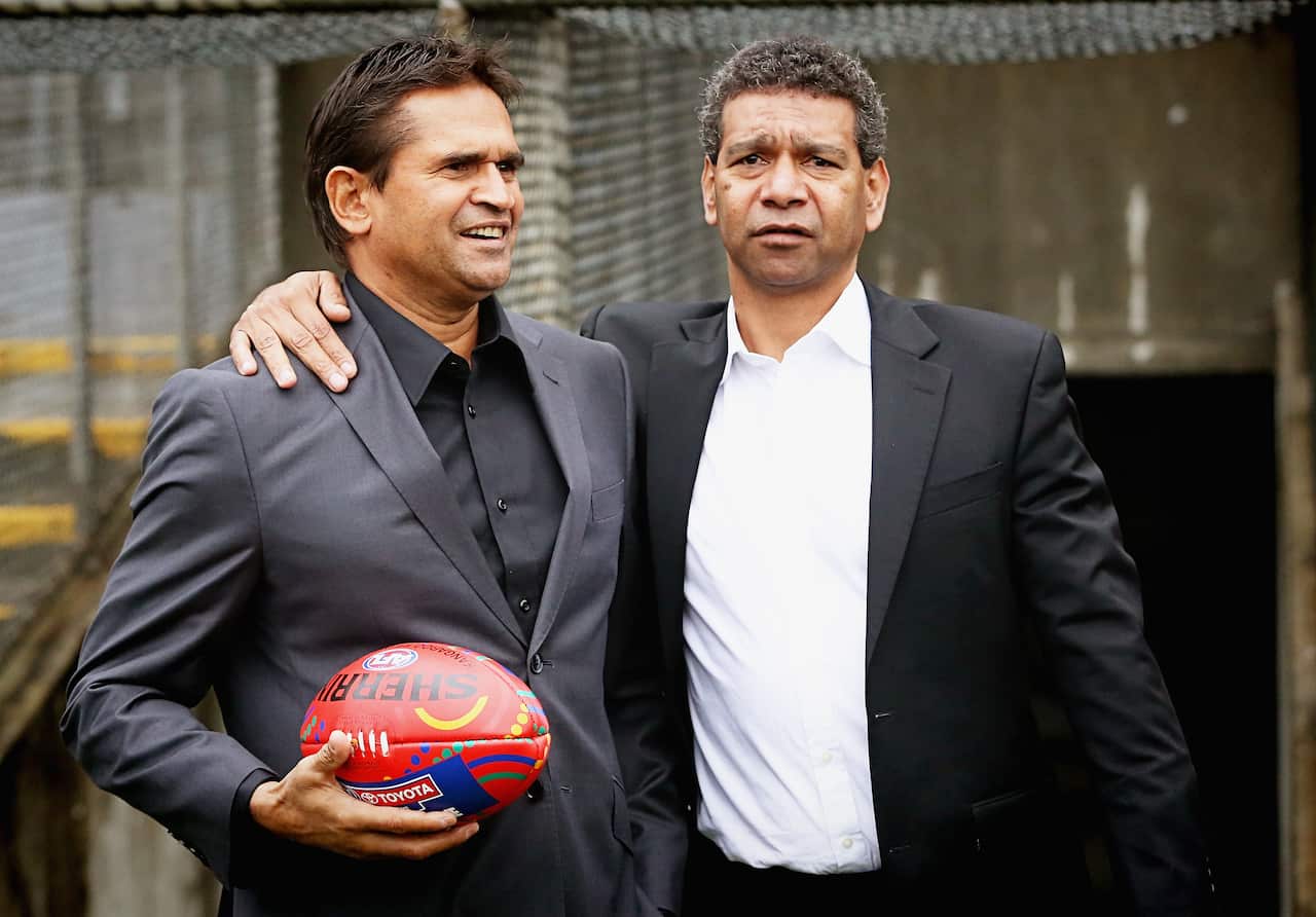 Two mean wearing formal attire standing next to each other. One is holding an Aussie rules football.