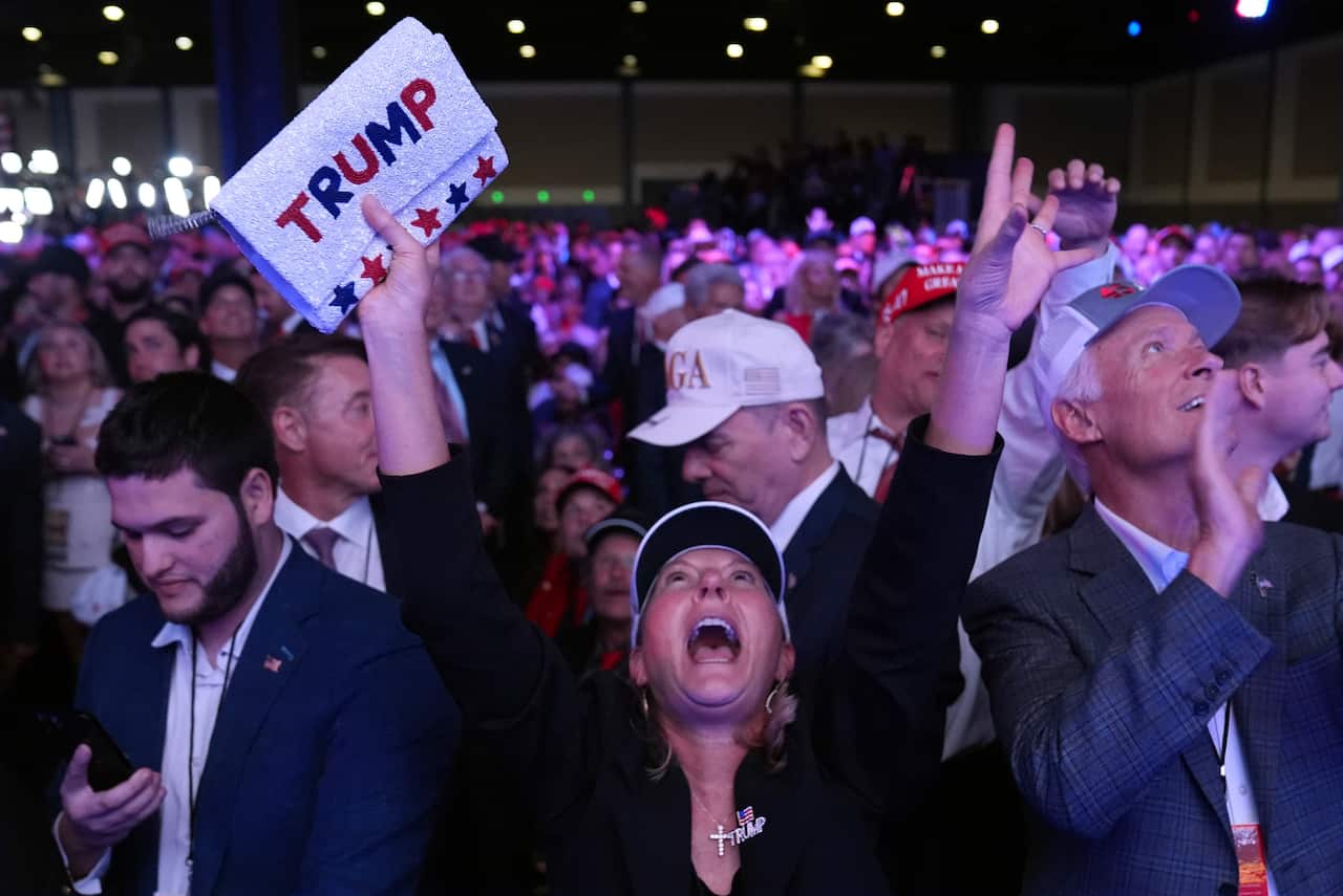 Jubilant supporters celebrate at a campaign election night watch party for Republican presidential nominee Donald Trump. In the centre foreground, a woman shouts with her arms aloft. A towel with "Trump" emblazoned on it is in her right hand.