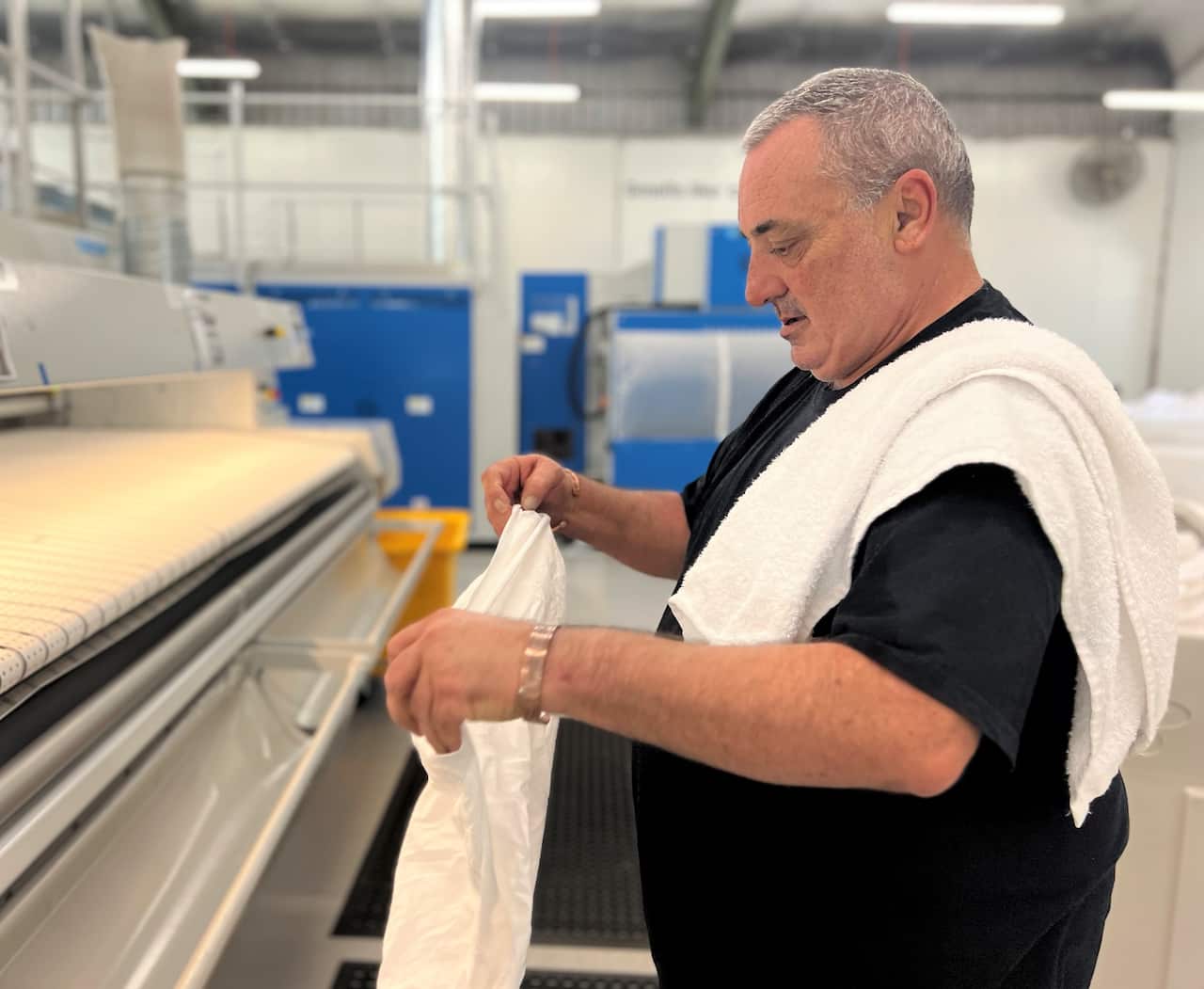 A man in a black t-shirt stands at an ironing machine folding linen.