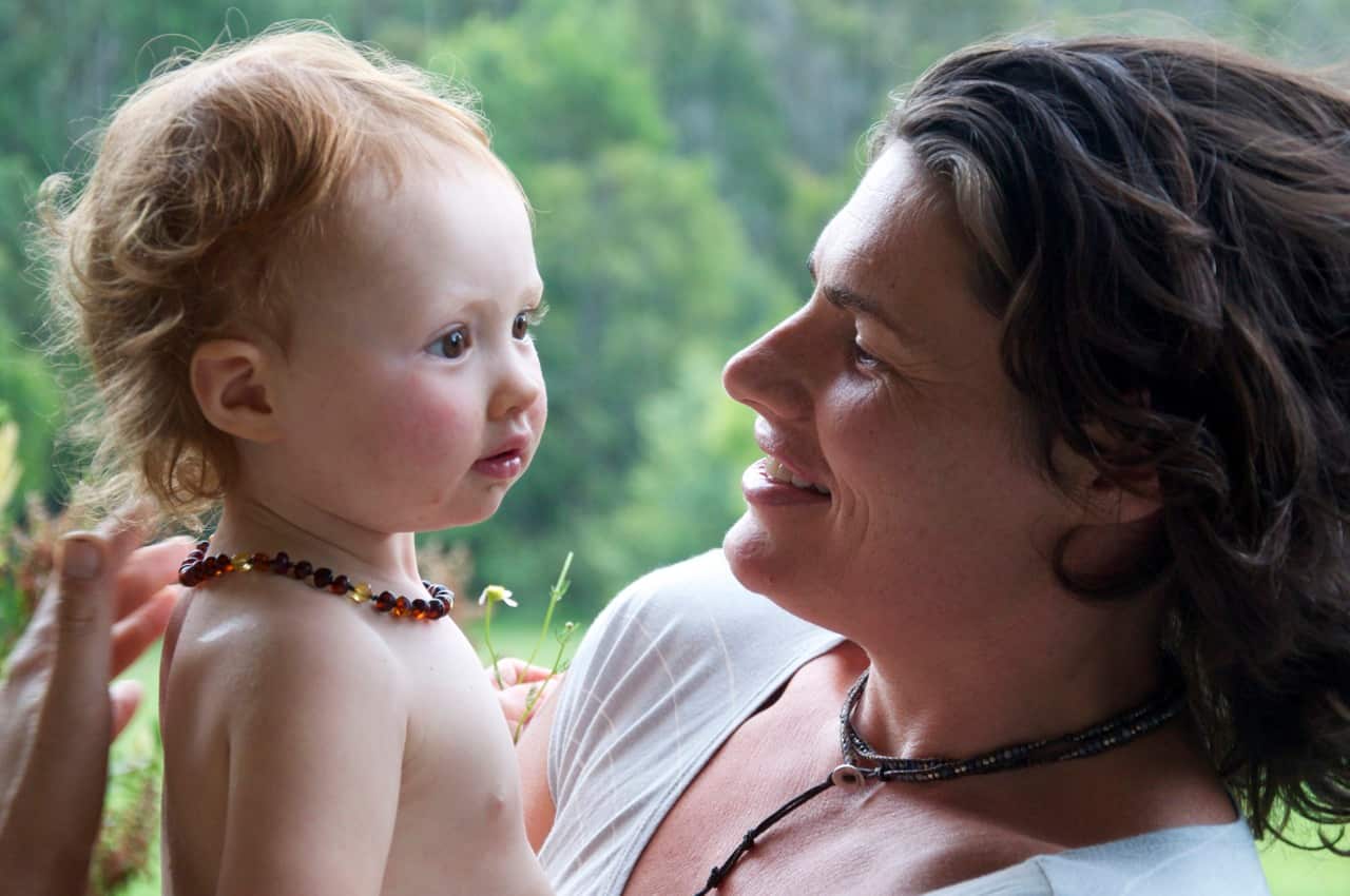 A mother smiles as she cuddles her daughter, who is bare-chested and wearing a necklace.