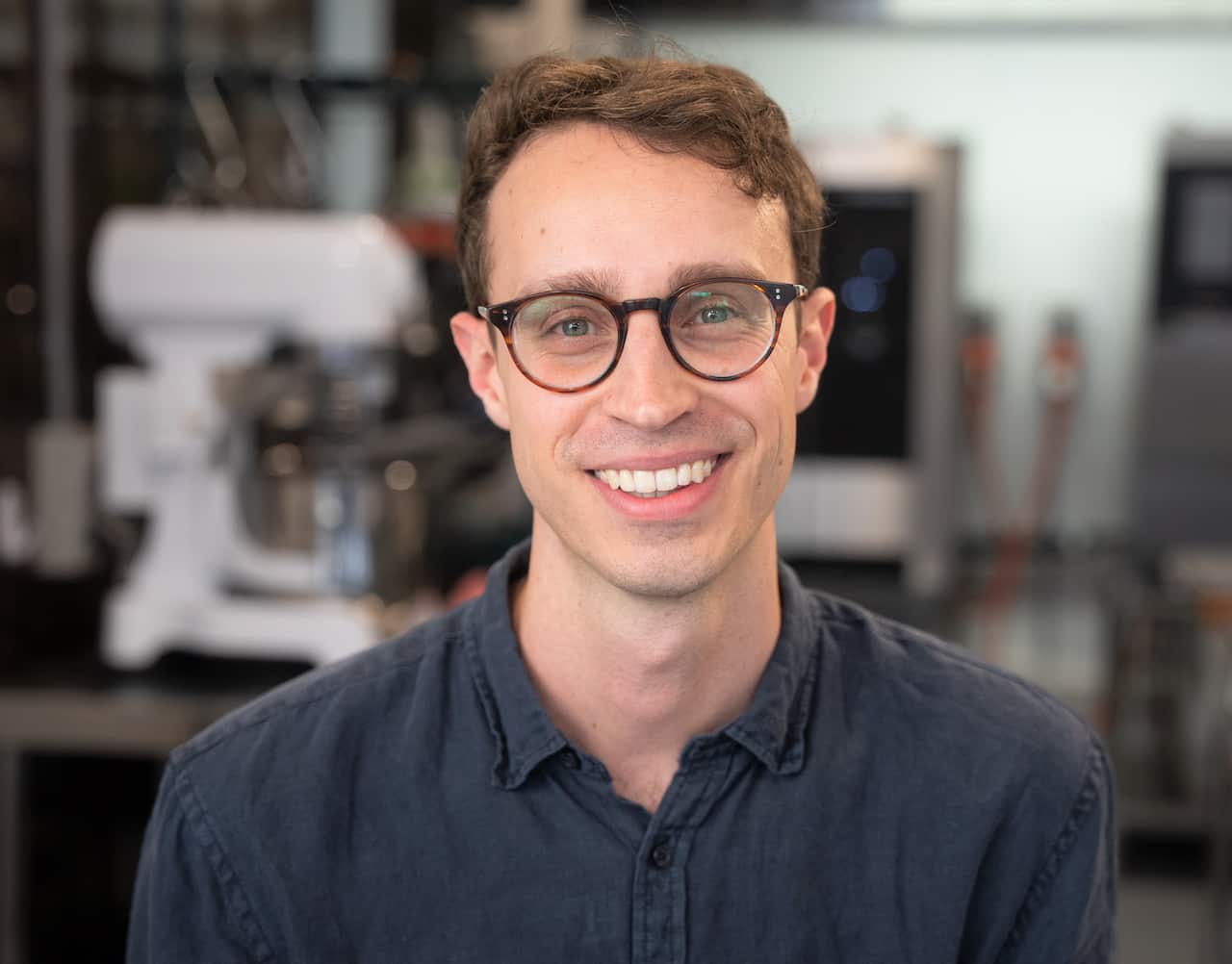 A man in a grey shirt sits on a stool smiling at camera.