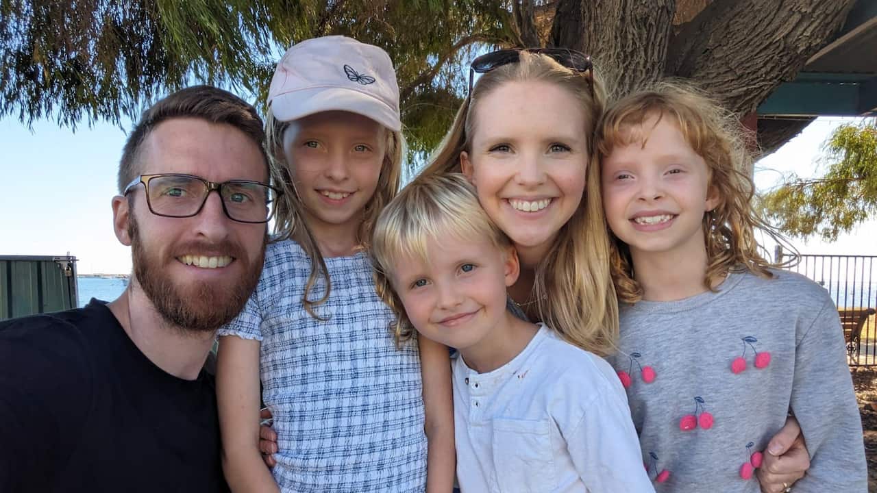 A family poses for a photograph while standing close together outside. From left, a man with beard and glasses, a young girl wearing a white cap, a young boy with blond hair, a woman with long blonde hair and a young girl with long red hair.