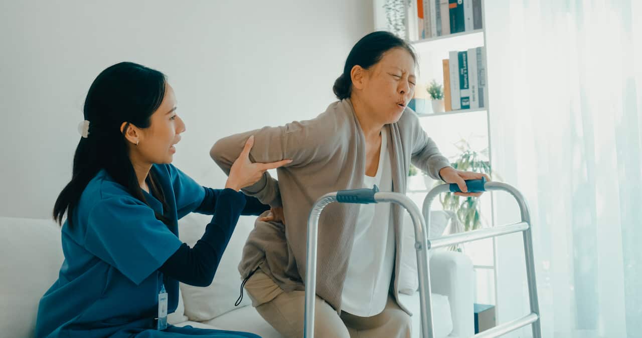 Young Asian nurse helps an elderly woman experiencing back pain use walker, support and care in living room at home. Home health care.