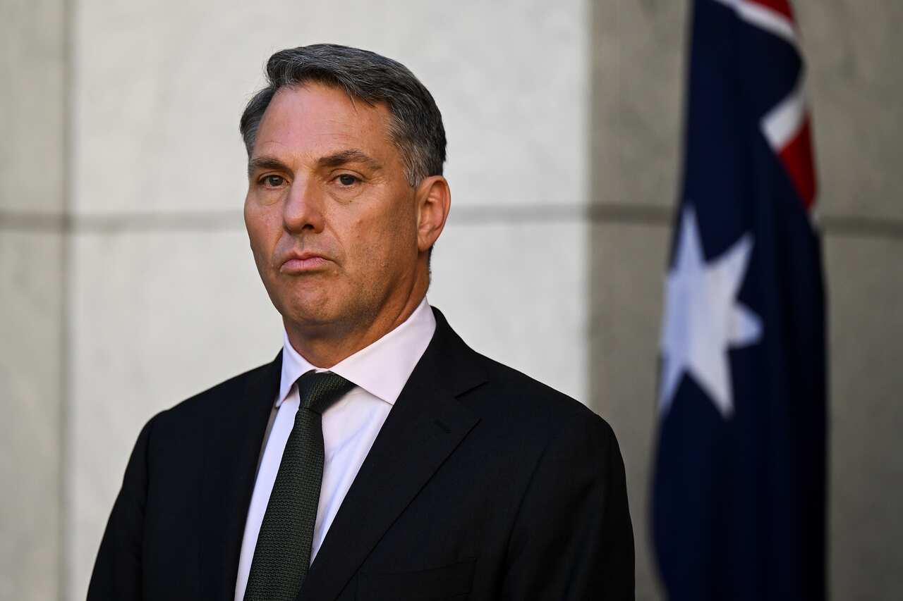 Richard Marles wearing a dark suit and standing outside. There is an Australian flag behind him.