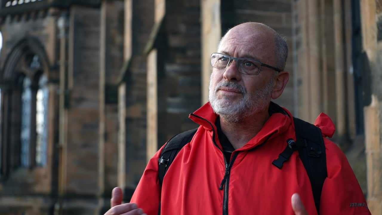 Tim Flannery is red parka, standing outside a sandstone building. 