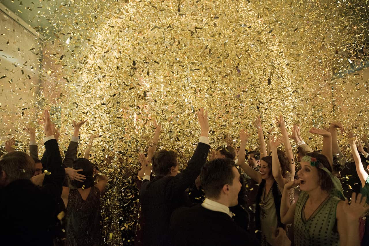 A dancing, smiling crowd, including women in flapper dresses, cheer with arms in the air, under a cascade of golden glittery pieces.