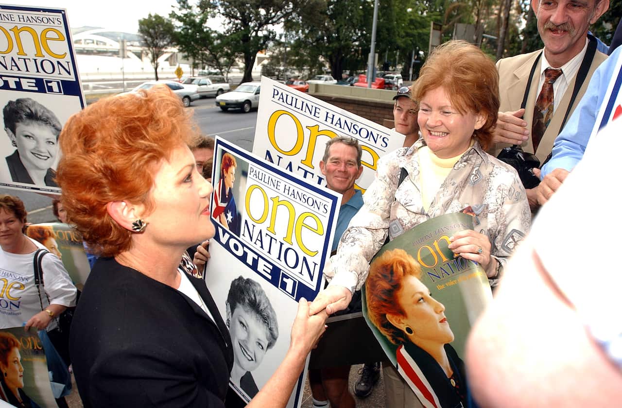 Pauline Hanson shakes hands with a smiling supporter on a street campaign trail, surrounded by people holding "Pauline Hanson's One Nation" election campaign signs and posters.