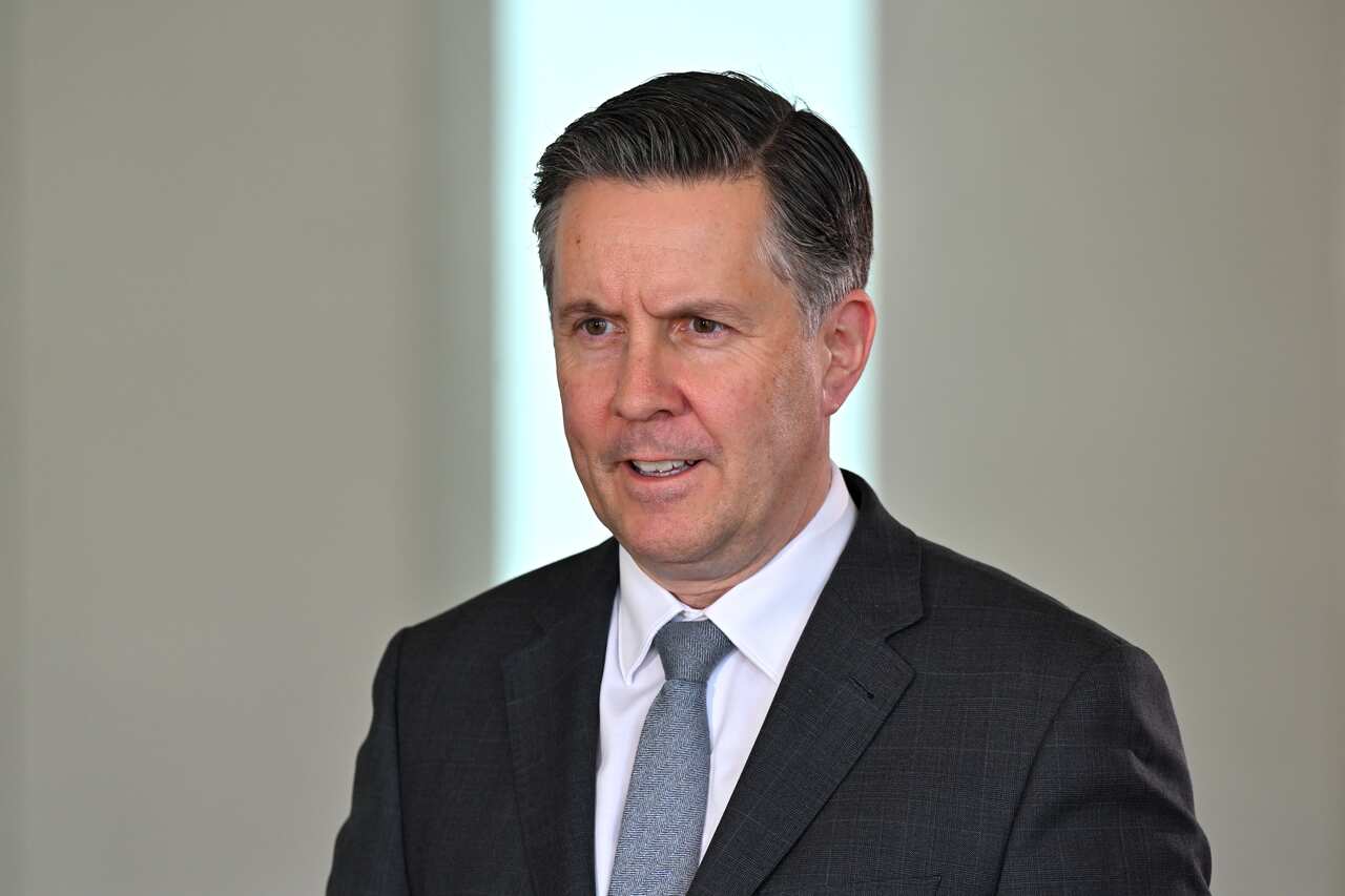 A man in a dark grey suit, a white shirt and blue tie, speaking in front of a white wall.