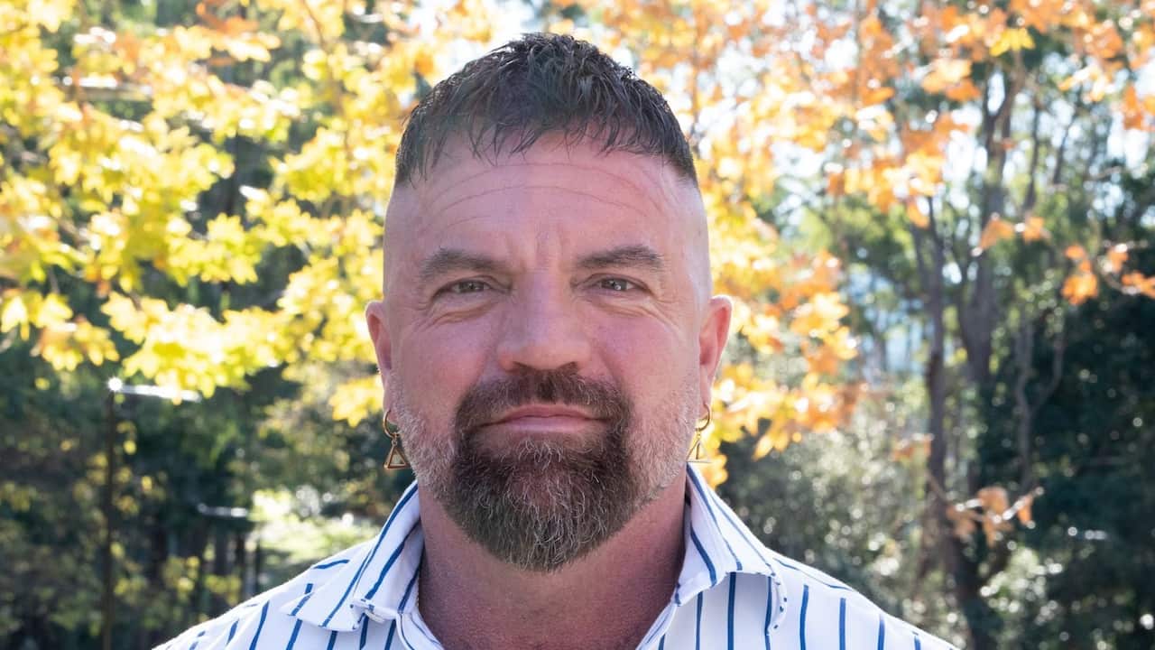 a middle aged man with a brown beard and hair in a stripey collared shirt smiles at the camera, autumn leaves are in the background