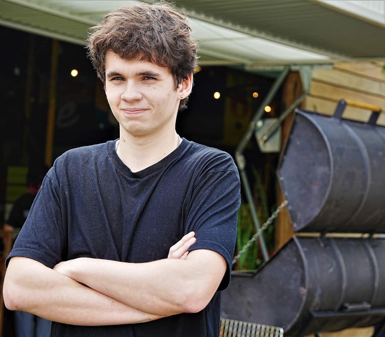 A young man in a black t-shirt stands with arms folded. 