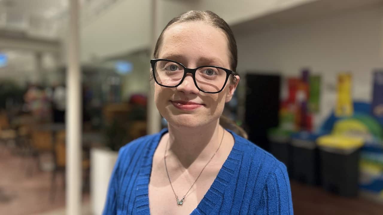 a young girl with wide-rimmed glasses and in a blue jumper smiles at the camera in a white office