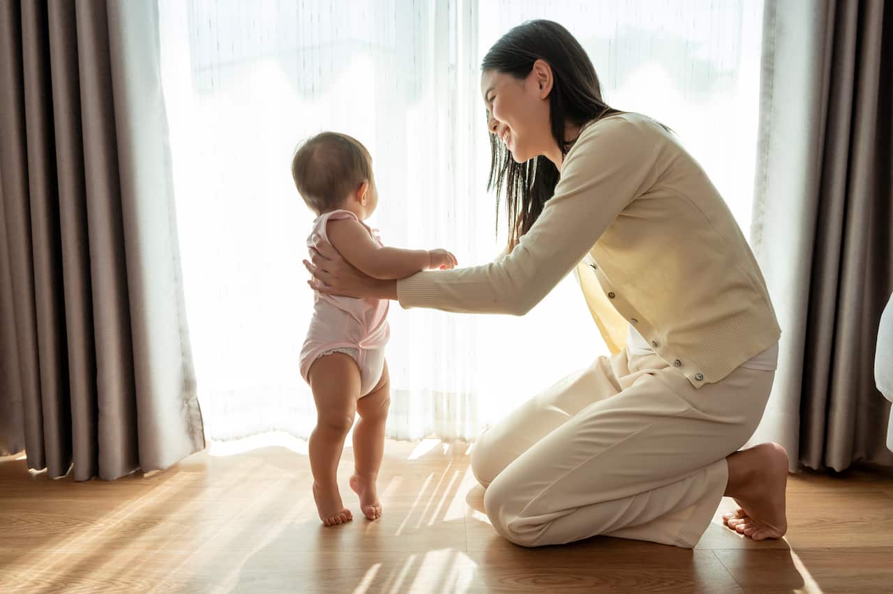 Baby girl stands on tippy toes while mother holds her.
