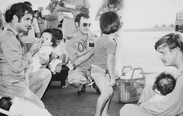 RAAF pilots holding Vietnamese babies, feeding them with bottles on an airport tarmac.