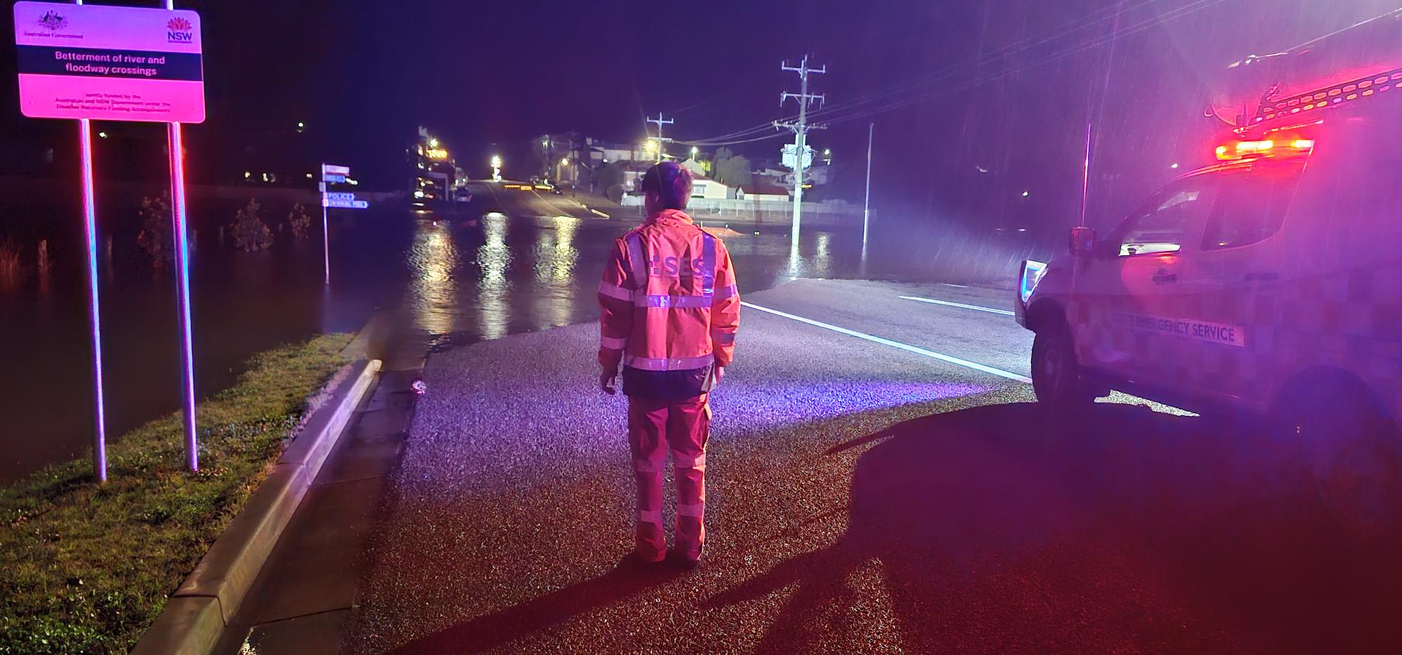 A NSW SES volunteer stands facing a flooded section of road at night time. 