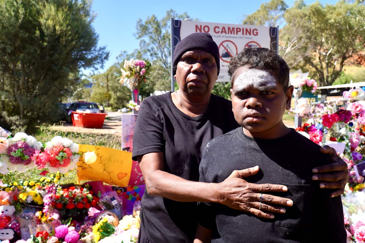 A woman stands behind a young man, both wearing black t-shirts. A large pile of flowers behind them.