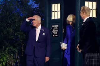 King Charles, wearing a dark blue suit and light tie, holds his right hand to his brow as though peering forward. Behind him is Queen Camilla in a blue dress and a man in a dark suit. Camilla is emerging from a large blue box with lighted windows.
