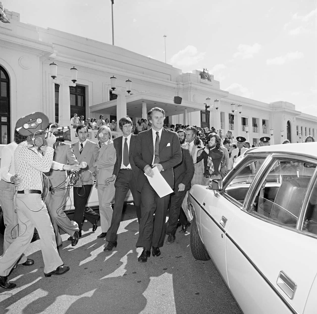 Black and white photo of then Opposition leader Malcolm Fraser walks away from old parliament house towards a parked car, with reporters and cameras following him.