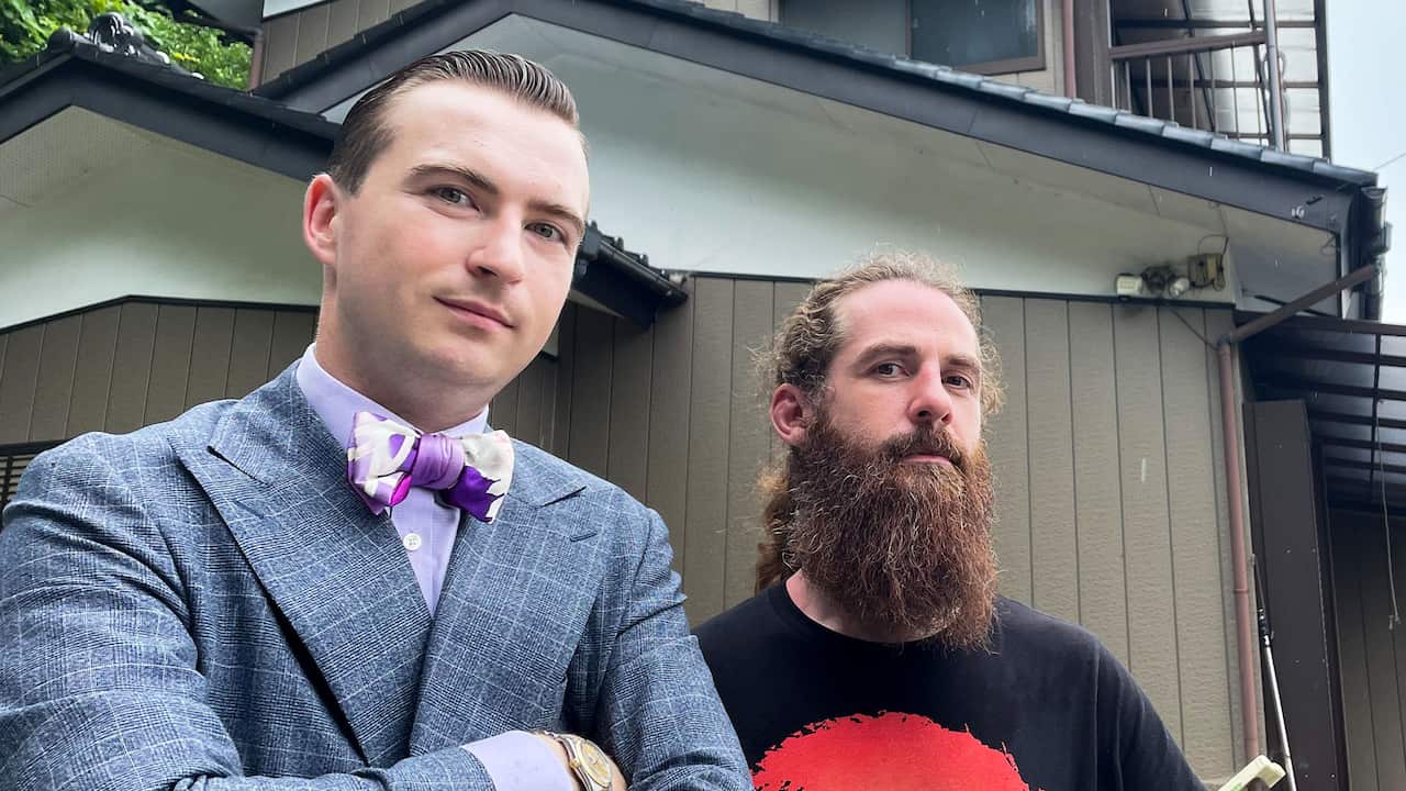 Two man pose for a photo. The man on the left is wearing a grey suit, lavender-coloured shirt and a light purple bow tie. The man on the right has a long beard and is wearing a black t-shirt