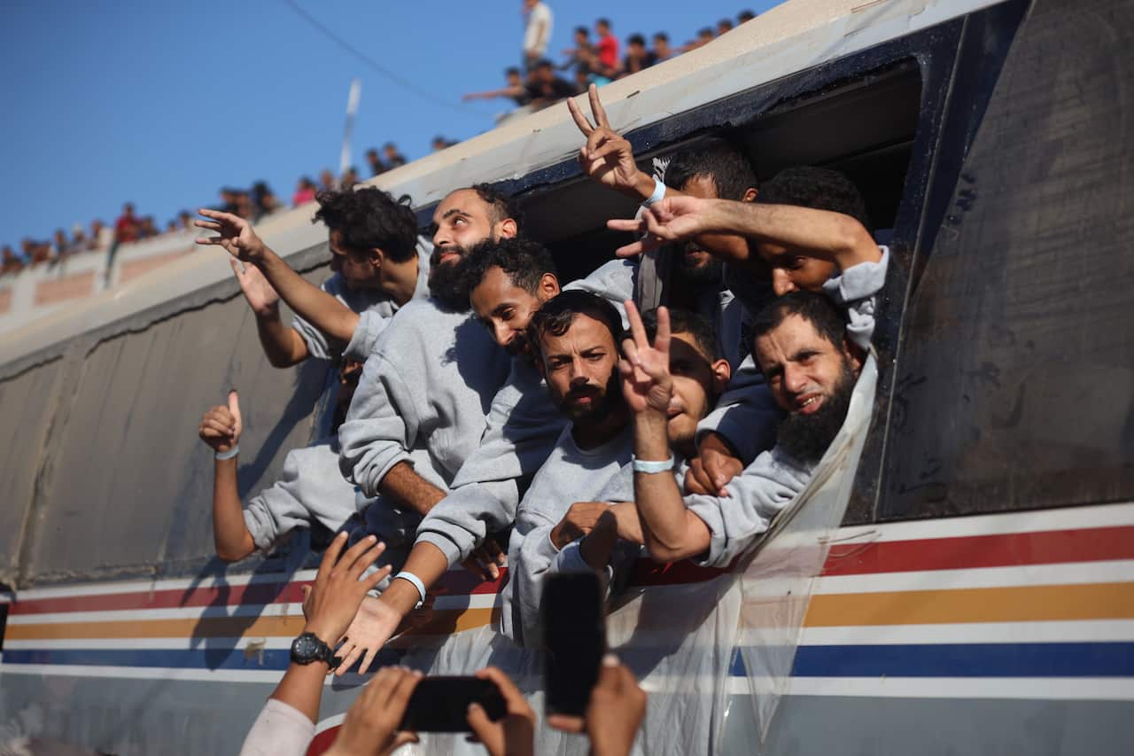 A group of Palestinian men in grey sweatshirts wave and show peace signs and thumbs up from the window of a bus.