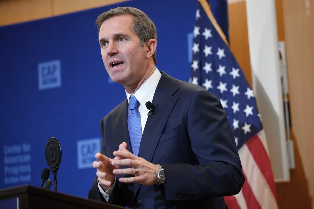 A man in a navy suit speaking inside in front of a blue sign and a US flag.