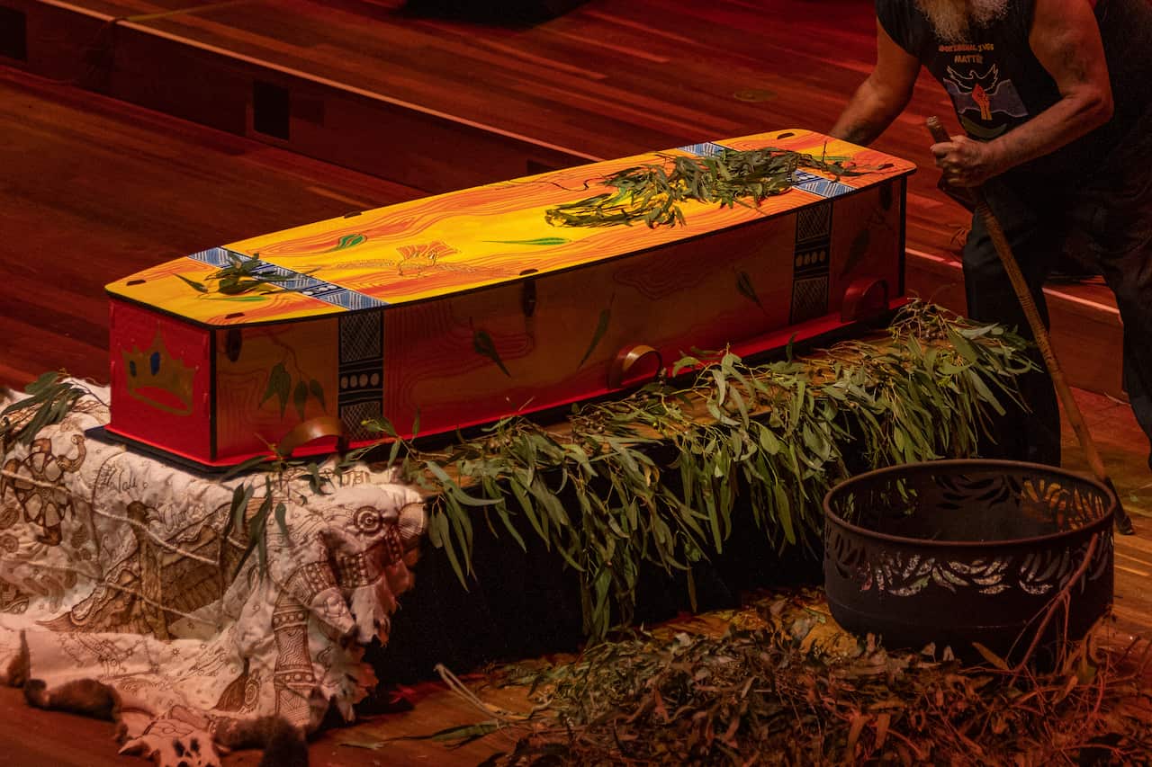 Uncle jack charles' red and yellow coffin sitting atop gum leaves and a possum skin cloak.