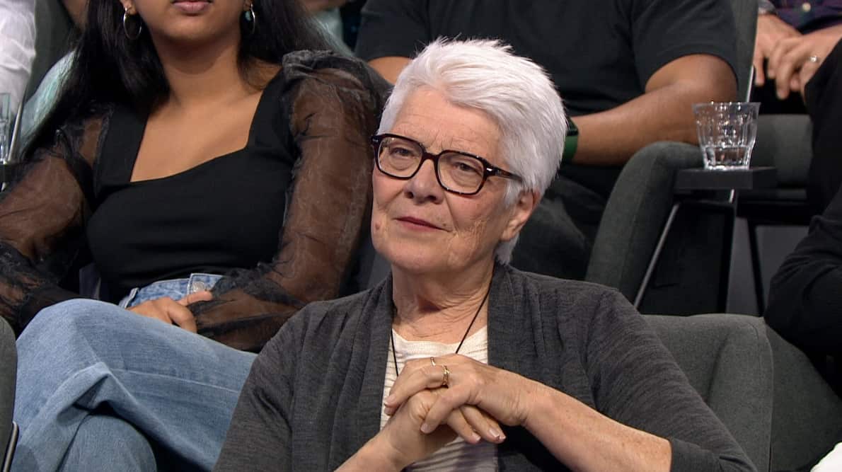 A woman with grey hair and glasses sitting in a TV studio.