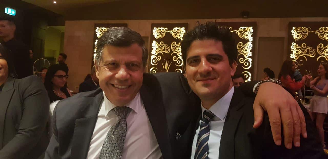 Two men in dark suits and ties are smiling while posing for a photo at a table. The man on the right has his arm around the man on the left, and the background shows other attendees in a warmly lit room with decorative wall panels.